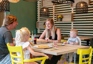 Gezin geniet samen van een maaltijd met pizza's aan een tafel in een gezellig ingericht restaurant op een vakantiepark.