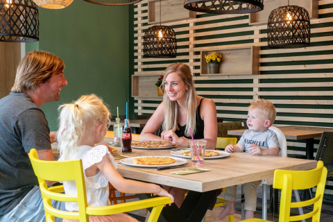 Gezin geniet samen van een maaltijd met pizza's aan een tafel in een gezellig ingericht restaurant op een vakantiepark.