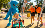 Børn leger på en legeplads med voksne og en stor blå maskot ved Aminess Style Camping Atea Resort i Kroatien.