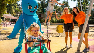 Børn leger på en legeplads med voksne og en stor blå maskot ved Aminess Style Camping Atea Resort i Kroatien.