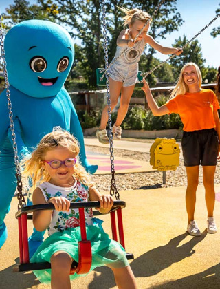 Børn leger på en legeplads med voksne og en stor blå maskot ved Aminess Style Camping Atea Resort i Kroatien.