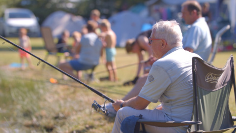 Oudere man vist ontspannen aan een vijver bij een vakantiepark met glamping, omringd door gezinnen.