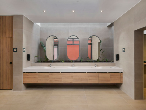 Salle de bains moderne d’un parc de vacances avec meubles en bois, miroirs, trois lavabos et quelques plantes vertes.
