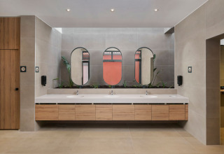 Baño moderno en un parque vacacional con gabinetes de madera, plantas y tres lavabos frente a espejos grandes.