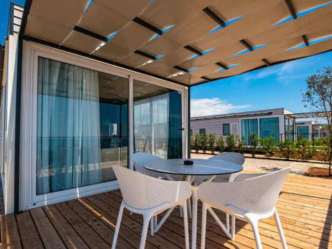 Modern chalet patio with round table and four chairs on wooden deck, shaded by a pergola on a sunny day.