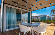 Moderne Chalet-Terrasse mit Holzboden, Tisch und vier Stühlen vor großen Glasschiebetüren bei Sonnenschein.