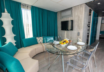 Moderne chaletstue med runde sofaer, turkise gardiner, glasbord og gennemsigtige stole, klar til spisning.