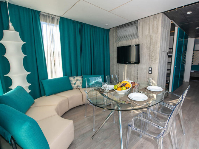 Salotto di chalet moderno con divano curvo, tende turchesi, tavolo in vetro e sedie trasparenti apparecchiate.