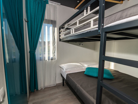 Modern chalet room with bunk beds, teal curtains, large window, and sunlight streaming onto wood floors.