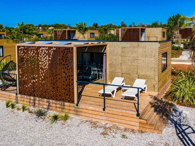 Moderne chalet kaldet Camping Home Green med træterrasse og liggestole under klar blå himmel.