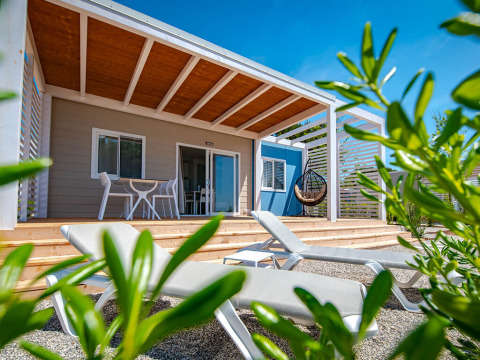 Chalet mit Holzterrasse, Sonnenliegen und Hängesessel, umgeben von Pflanzen und strahlend blauem Himmel.