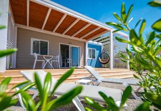 Chalet mit Holzterrasse, Sonnenliegen und Hängesessel, umgeben von Pflanzen und strahlend blauem Himmel.
