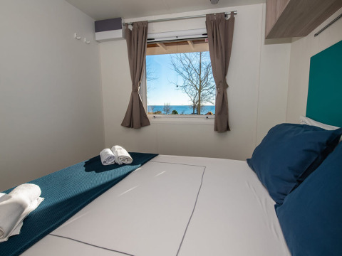 Chalet-Schlafzimmer mit blauen Kissen, Handtüchern und Fensterblick auf das Meer und Bäume draußen.