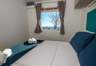 Chalet-Schlafzimmer mit blauen Kissen, Handtüchern und Fensterblick auf das Meer und Bäume draußen.