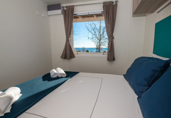 Soveværelse i chalet med blå puder og håndklæder, vindue med udsigt over havet og træer udenfor.