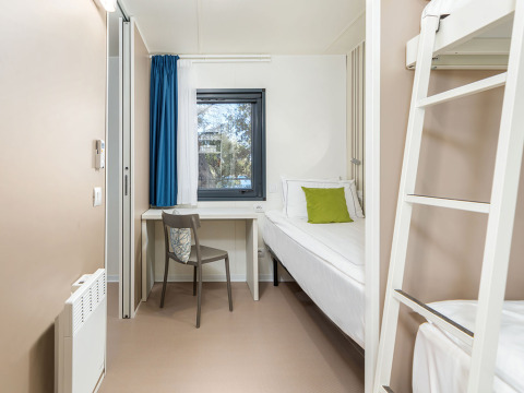 Helles Zimmer in der Camping Villa mit Einzelbett, Etagenbett, Schreibtisch und Fenster mit blauen Vorhängen.
