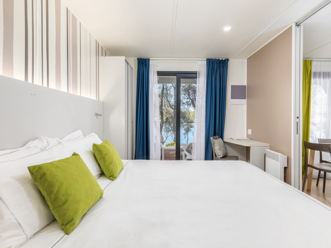 Modern camping villa bedroom with white bed, green pillows, desk, and sea view through glass doors.