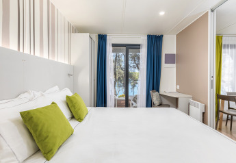 Modernes Camping-Villa-Schlafzimmer mit weißem Bett, grünen Kissen und Meeresblick durch Glastüren.