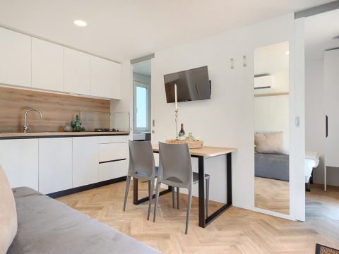 Bright, modern lodge interior with compact kitchen, dining table set for two, and a view into the bedroom.