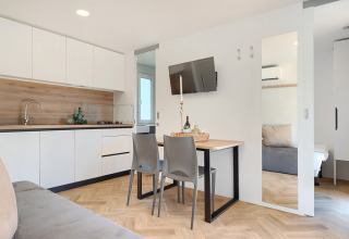 Bright, modern lodge interior with compact kitchen, dining table set for two, and a view into the bedroom.