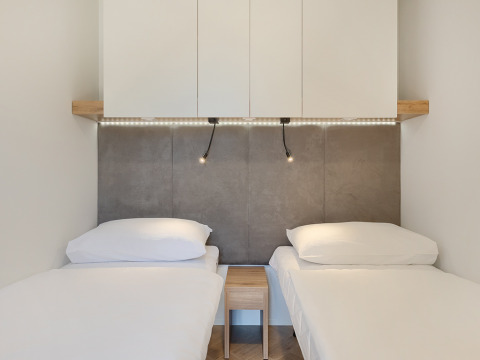 Two single beds with white linens, a nightstand, wall panel, overhead cabinets, and LED lights in a lodge room.