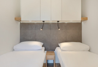 Deux lits simples avec draps blancs, table de nuit, panneau mural, armoires en hauteur et éclairage LED dans un lodge.