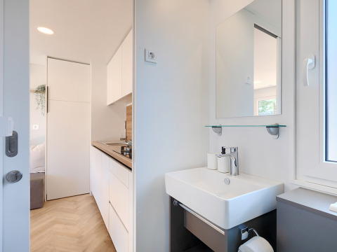 Modern lodge interior showing a small bathroom sink, mirror, and adjacent minimalist kitchen area.