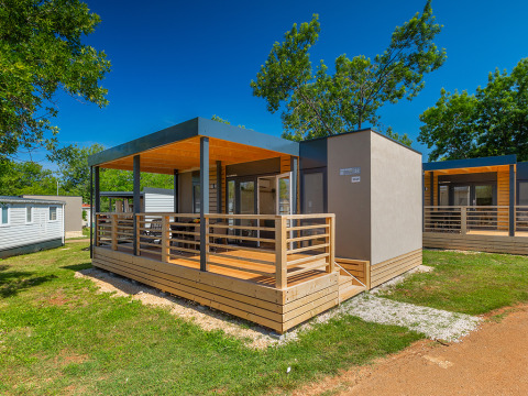 Lodge moderne avec terrasse en bois, entourée d’arbres et de pelouse sous un ciel bleu éclatant.