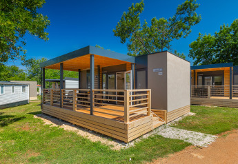 Lodge moderne avec terrasse en bois, entourée d’arbres et de pelouse sous un ciel bleu éclatant.
