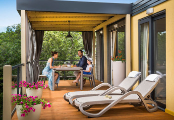 Familie nyder morgenmad på terrassen af Bella Vista Home-lodge med udsigt over grønne træer og blomster.