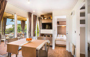 Interior de lodge moderno con comedor, dormitorio abierto, balcón, grandes ventanales y cortinas marrones.
