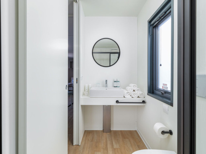 Modernes Badezimmer im Camping Home Sea View mit Waschbecken, kreisförmigem Spiegel und frischen Handtüchern.