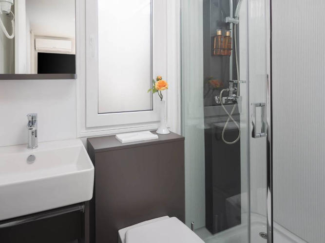 Bagno moderno con doccia in vetro, lavabo quadrato, finestra opaca e fiore giallo all’Aminess Style Camping Avalona Resort.