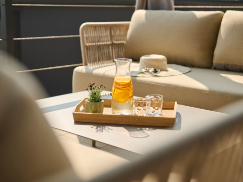 Kande med citronvand, to glas og en lille plante på bakke, placeret på bord ved komfortabel udendørs sofa.