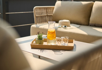 Kande med citronvand, to glas og en lille plante på bakke, placeret på bord ved komfortabel udendørs sofa.