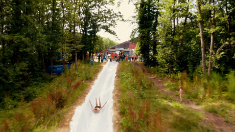 A person slides down a long water slide at Camping Fuussekaul in Wiltz, Luxembourg, surrounded by trees.