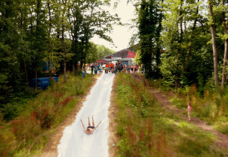 A person slides down a long water slide at Camping Fuussekaul in Wiltz, Luxembourg, surrounded by trees.