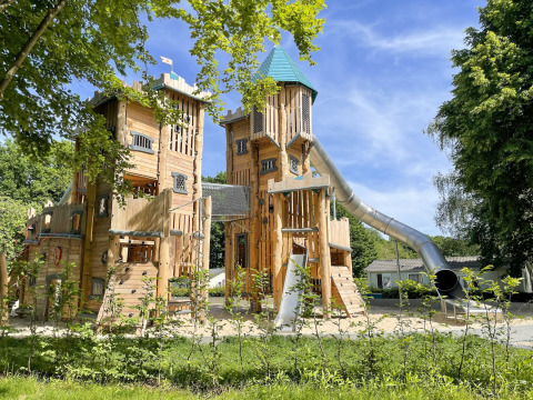 Large wooden playground castle with high towers and a slide at Camping Fuussekaul in Wiltz, Luxembourg.