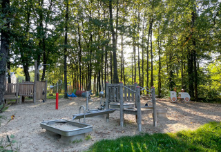 Playground surrounded by trees at Camping Fuussekaul in Wiltz, Luxembourg, featuring sand and play equipment.