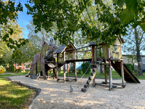 Wooden playground with slides and climbing towers on sand, surrounded by green trees on a sunny day.