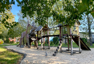 Wooden playground with slides and climbing towers on sand, surrounded by green trees on a sunny day.