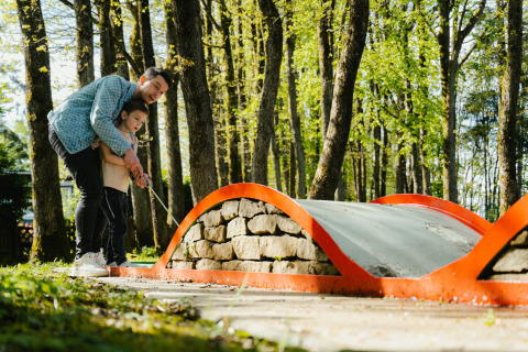 An adult helps a child play mini golf on a wooded course at Camping Fuussekaul in Wiltz, Luxembourg.