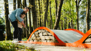 Een volwassene helpt een kind bij minigolf spelen in een bosrijk park bij Camping Fuussekaul, Wiltz.
