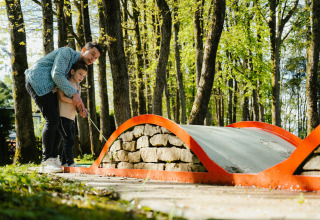 Un adulto ayuda a un niño a jugar al minigolf en un parque arbolado en Camping Fuussekaul en Wiltz.