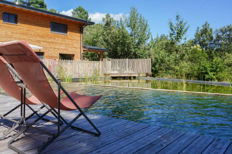 Liegestühle an einem Naturpool neben einem Holzhaus im Ferienpark Bivouac nature in Okzitanien, Frankreich.