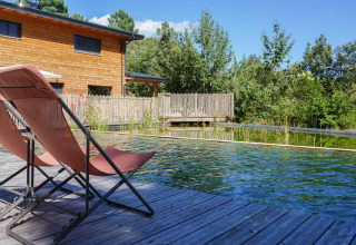 Transats au bord d'une piscine naturelle et maison en bois au parc de vacances Bivouac nature en Occitanie, France.