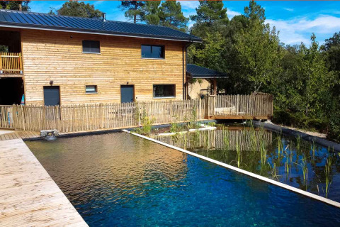 Natural swimming pool and wooden building at Bivouac Nature holiday park in Occitanie, France, among trees.