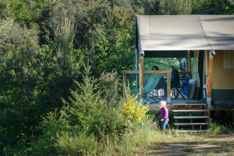 A cabin at Bivouac Nature holiday park in Occitanie, France, with people relaxing on the porch and a child outside.