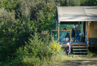Cabane au Bivouac Nature, parc de vacances en Occitanie, France, avec des personnes sur la terrasse et un enfant dehors.