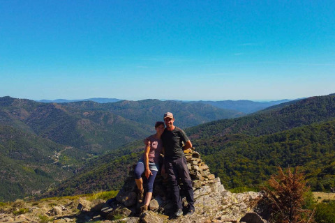 A couple stands on a rocky mountain viewpoint at Bivouac nature holiday park in Occitanie, France, amid green hills.
